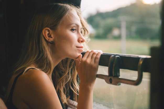 Estado de ánimo atmosférico estilo de vida retrato de la joven y bella chica de cabello rubio mirando por la ventana del tren. Bastante adolescente disfrutando de la belleza de la naturaleza del vagón de tren en movimiento en verano.