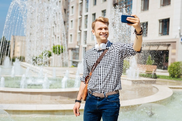 Estado de ánimo placentero. Hombre guapo feliz sosteniendo su teléfono inteligente mientras toma un selfie