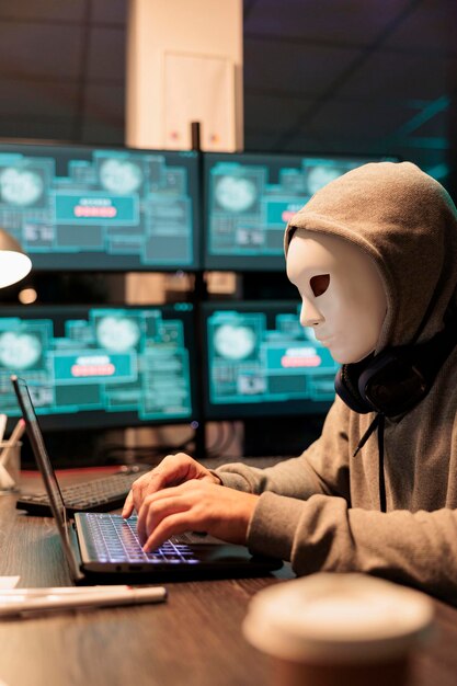 Estafador masculino aterrador que usa máscaras que piratean servidores de bases de datos, hacker con capucha irrumpiendo en el sistema informático y activando virus para crear malware. Misterioso impostor robando grandes datos.