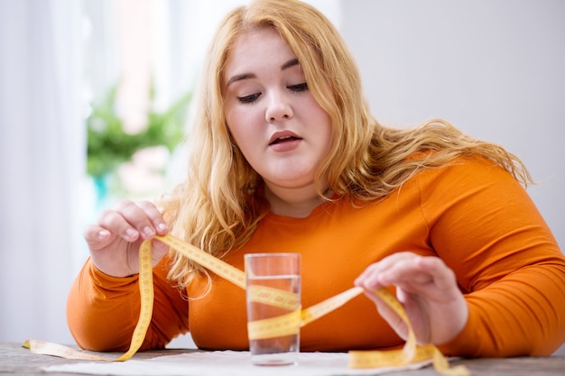 Estar acima do peso. mulher concentrada e obesa sentada à mesa e segurando uma fita métrica