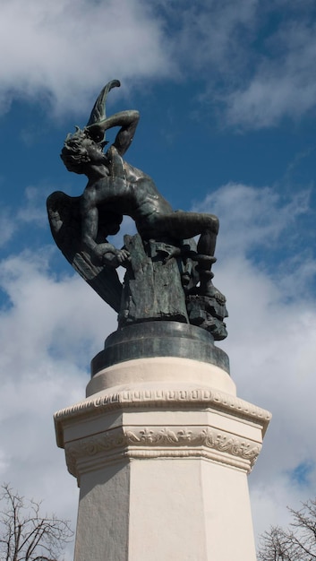 Estatua de una persona que representa al Ángel Caído en negro con alas y una serpiente sobre un pedestal de granito gris en el Parque del Retiro de Madrid