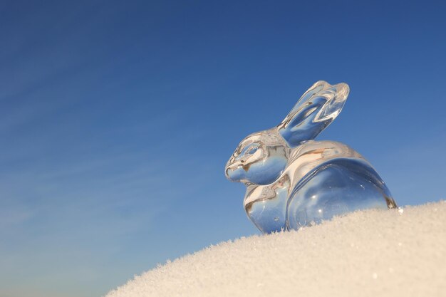 Estatueta de coelho transparente na neve branca contra um cartão de ano novo de fundo azul do céu