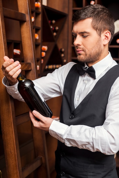 Este vinho é perfeito. Sommelier masculino confiante examinando uma garrafa de vinho em pé perto da prateleira de vinho