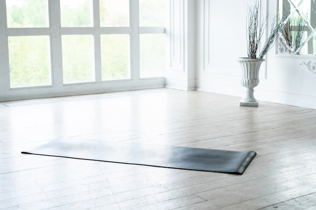 Estera de yoga desenrollada acostada en un estudio de fitness ligero vacío en un piso de madera, equipo deportivo desplegado preparado para el entrenamiento, sala de loft de pilates moderna lista para la sesión de entrenamiento. Concepto de vida saludable.