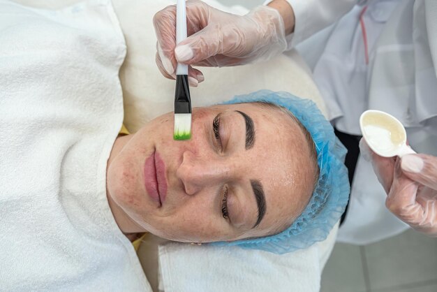 Esteticista aplica una máscara en la cara del paciente con cepillos cosméticos mientras está acostado