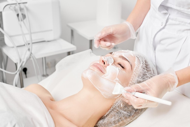 Esteticista aplica máscara de crema en la cara de la mujer para el procedimiento de rejuvenecimiento de la piel de la cara en el salón de belleza