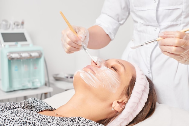Esteticista aplicando máscara de crema cosmética en la cara de la mujer para el procedimiento de rejuvenecimiento en el salón de belleza