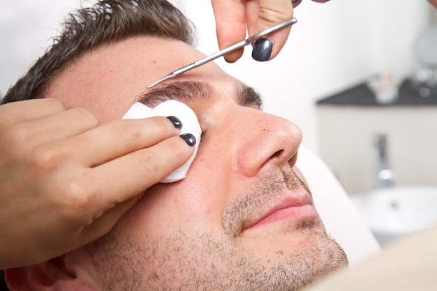 Foto esteticista arrancando as sobrancelhas de um homem bonito com uma pinça em um salão de beleza