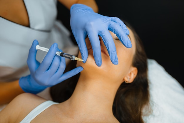 Esteticista de luvas aplica injeção de botox no queixo em paciente do sexo feminino na mesa de tratamento. Procedimento de rejuvenescimento em salão de esteticista. Médico com seringa e mulher, cirurgia estética contra rugas