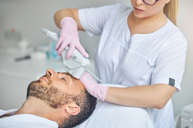 Esteticista feminina em luvas estéreis tratando a pele masculina com dispositivo a laser