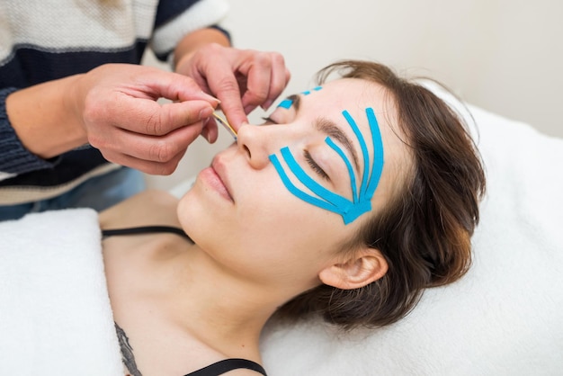 Esteticista feminino fazendo a gravação do rosto do paciente deitado na mesa no escritórioEspecialista realizando aplicação de kinesio taping no rosto da menina