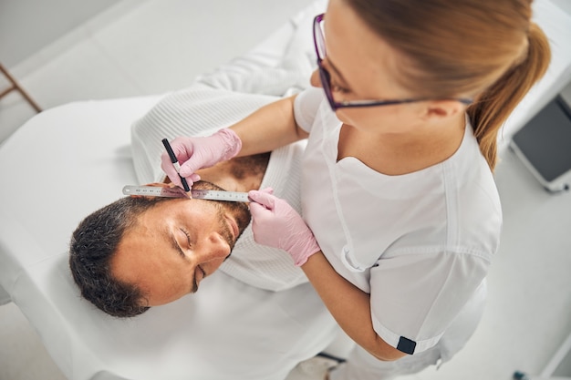 Esteticista en guantes estériles colocando la regla en la mejilla del joven y dibujando una línea recta con lápiz blanco