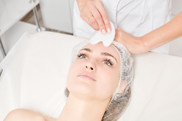 Esteticista haciendo masaje facial con piedra Gua Sha de la piel de la cara de la mujer para el drenaje linfático