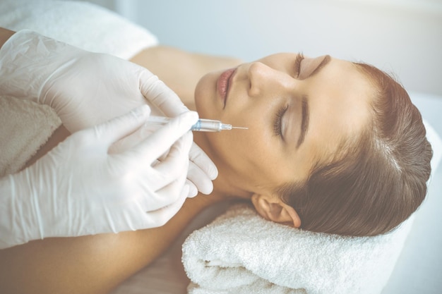 Esteticista haciendo un procedimiento de belleza con una jeringa en la cara de una joven morena en una clínica soleada. Medicina y cirugía estética, inyecciones de belleza.