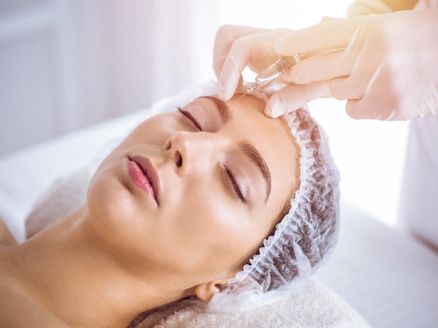 Esteticista haciendo un procedimiento de belleza con una jeringa en la cara de una joven morena en un soleado centro de spa. Medicina y cirugía estética, inyecciones de belleza.