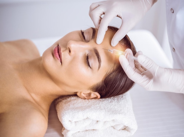 Esteticista haciendo un procedimiento de belleza con una jeringa en la cara de una joven morena en un soleado centro de spa. Medicina y cirugía estética, inyecciones de belleza.