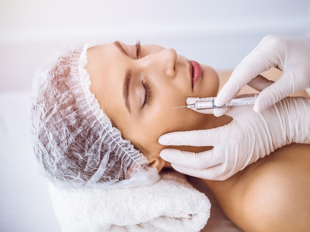 Esteticista haciendo un procedimiento de belleza con una jeringa en la cara de una joven morena en un soleado centro de spa. Medicina y cirugía estética, inyecciones de belleza.