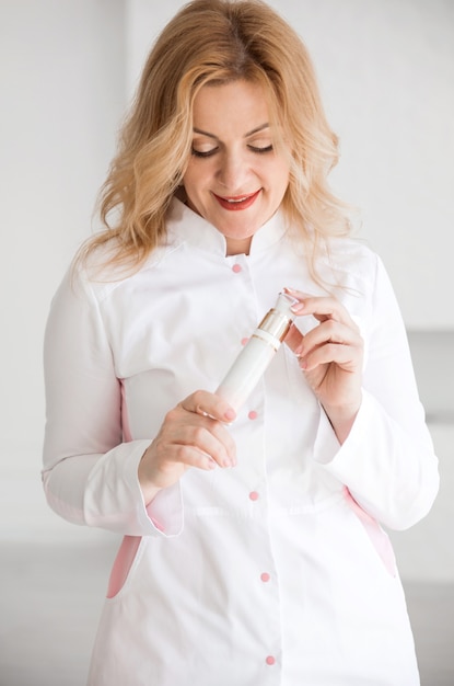 Esteticista hermosa chica en un traje blanco posando contra una pared blanca con un producto cosmético en sus manos.