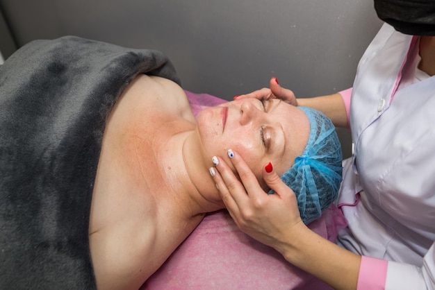 La esteticista masajea la cara y la cabeza de una mujer adulta acostada sobre una camilla de masaje. El concepto de cosmetología, procedimientos de spa de masajes.