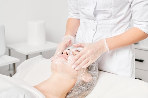 Esteticista masajea la máscara de crema en la piel de la cara femenina para el procedimiento de rejuvenecimiento en el salón de belleza