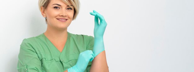 Esteticista médico se pone guantes azules estériles sonriendo se prepara para recibir clientes en blanco.