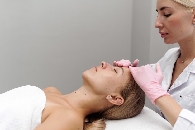 Esteticista o masajista haciendo manual relajante masaje rejuvenecedor de cara y cuello para mujer joven en salón de belleza. Masaje facial hidratante rejuvenecedor en cosmetología.