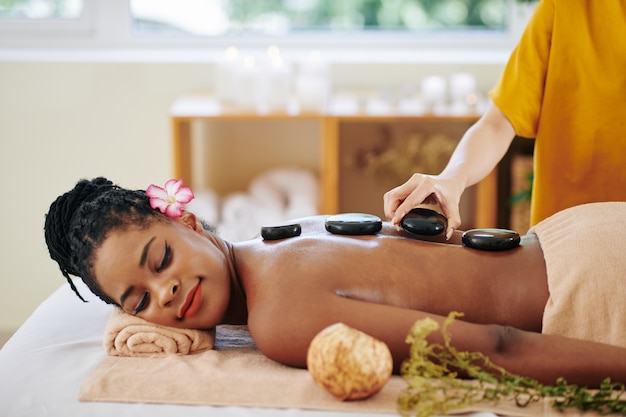 Esteticista poniendo piedras de basalto calientes a lo largo de la columna vertebral de una mujer negra bastante joven relajada