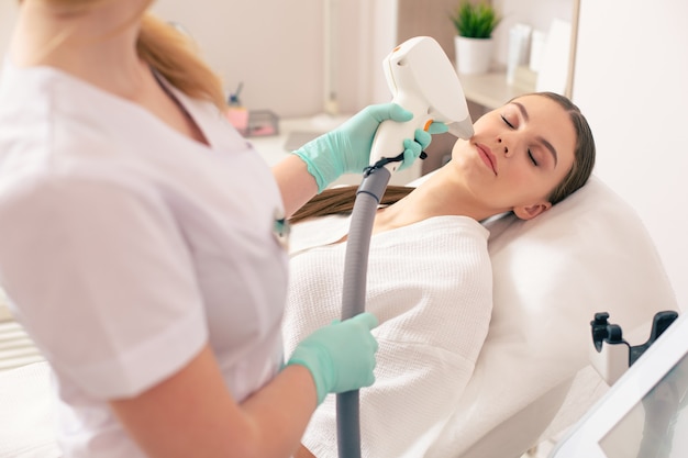 Esteticista profissional usando luvas de borracha e segurando um dispositivo moderno enquanto realiza o procedimento de depilação a laser com sua cliente