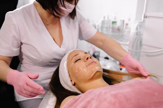 Esteticista remove uma máscara de argila do rosto de uma jovem no salão de spa Linda jovem na esteticista faz os procedimentos de spa