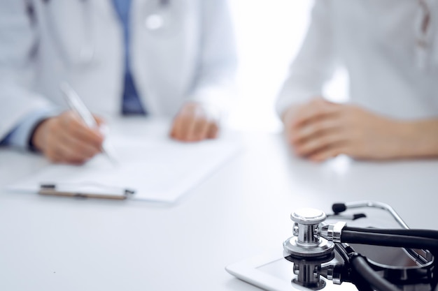 Estetoscópio deitado no computador tablet na frente de um médico e um paciente sentados um ao lado do outro ao fundo. Medicina, conceito de saúde.