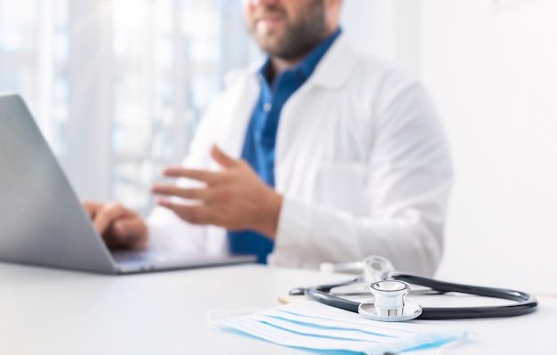 Foto estetoscópio e máscara médica na mesa dos médicos em segundo plano. médico realiza uma consulta ao paciente online usando o computador