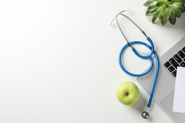 Estetoscópio, maçã, laptop e planta sobre fundo branco, vista superior. Local de trabalho médico