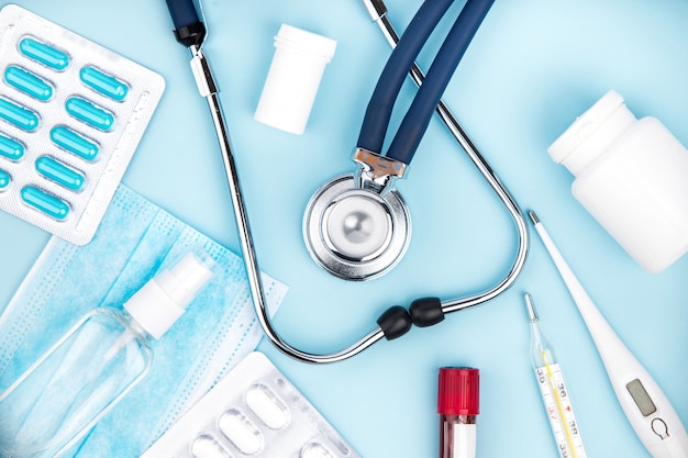 Estetoscópio, máscara médica, pílulas e termômetro sobre fundo azul.