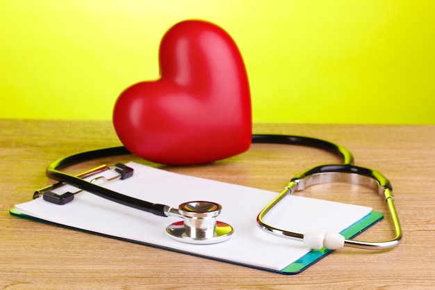 Estetoscopio médico con portapapeles y corazón sobre mesa de madera sobre fondo verde