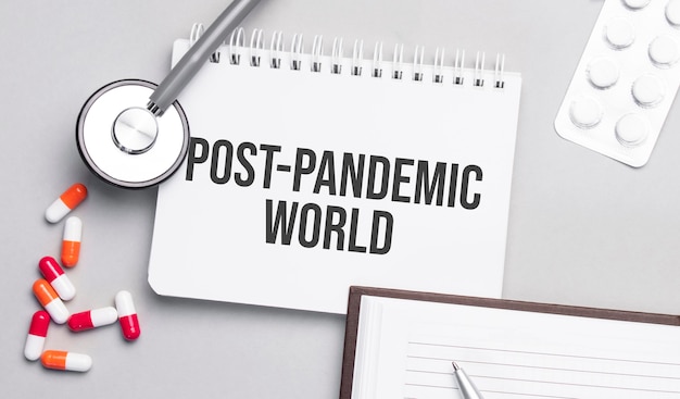Estetoscopio, píldoras y cuaderno con texto mundial pospandémico en la mesa médica