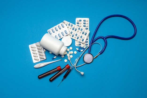 Estetoscópio, seringa, termômetro, tubo de ensaio de sangue e lata de comprimidos na vista superior de fundo azul com espaço de cópia.