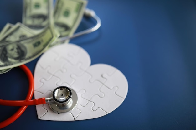 Un estetoscopio sobre el dinero y un corazón de rompecabezas Seguro médico Salud y medicamentos Cuánto cuesta el tratamiento Dinero y medicamentos Día Mundial del Corazón Día Mundial de la Salud Cuánto cuesta un corazón de donante