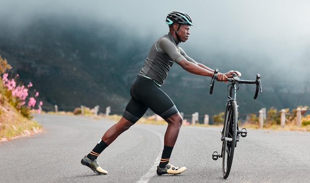 Esticando as pernas fitness e homem andando de bicicleta em uma estrada com uma bicicleta nas montanhas para viagens aeróbicas e exercícios esportivos na natureza Atleta fazendo um aquecimento antes de treinar em uma bicicleta na rua