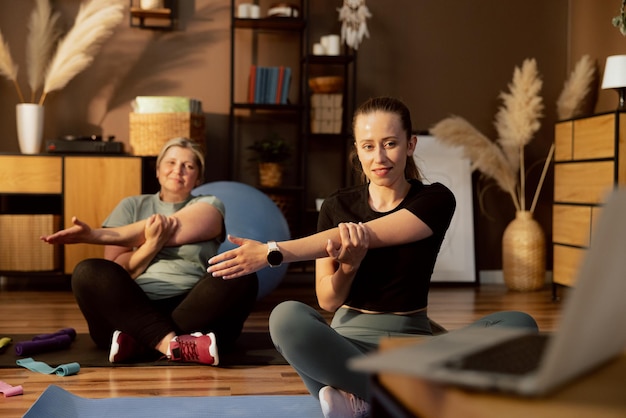 Esticando o tempo em casa mulher sênior com jovem sentada no tapete em pose de lotos assistindo aquecimento