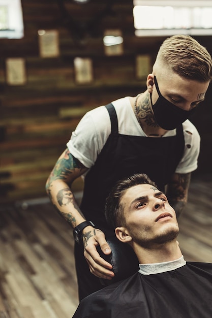 Estilista de cultivos preparando a un joven tatuado en una silla para afeitarse en una barbería