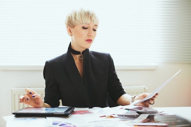 Estilista en elegante traje negro sentada a la mesa y leyendo, experta en moda. Mujer en el ámbito de la moda con tableta. Compras, adentro, cintura para arriba