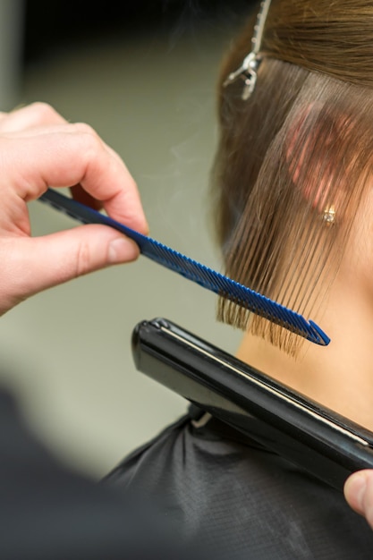 El estilista está alisando el pelo corto de una joven morena con una plancha plana en una peluquería de cerca