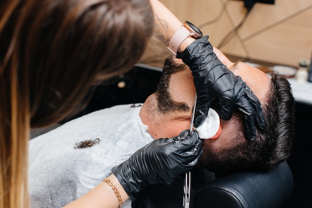 Un estilista profesional en una peluquería con estilo moderno afeita y corta el cabello de un joven.