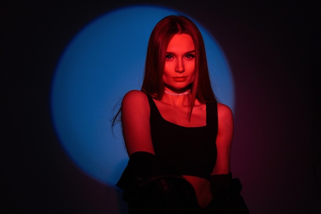 Estilizando a una joven hermosa con ropa de rock negra de moda con una chaqueta de cuero y un top de color rojo claro sobre una luz circular azul en el estudio sobre un fondo oscuro Retrato colorido