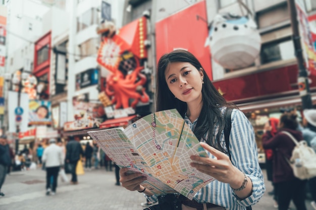 estilo antiguo de viaje autoguiado de jóvenes turistas asiáticos en osaka, japón, en vacaciones. elegante mujer viajera de pie en la calle dotonbori repleta de área popular sostener mapa de papel descubrir atracción turística