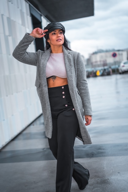 Estilo de la calle en la ciudad de una morena chica latina con un sombrero de cuero y un impermeable gris caminando en otoño