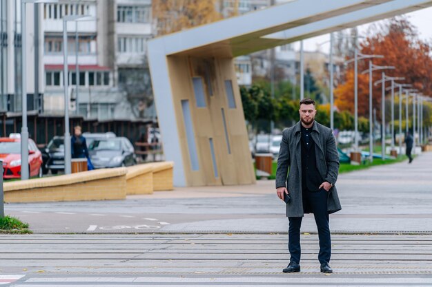 Estilo callejero. Hombre con un elegante traje negro y abrigo gris. Hombre caucásico joven hermoso que cruza una carretera central.