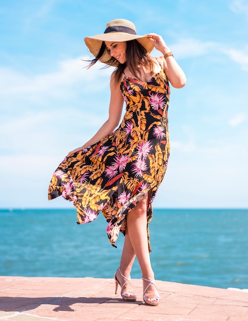 Estilo callejero, una joven morena con un vestido floral, un sombrero de paja y tacones. Vestido movido por el viento con el mar