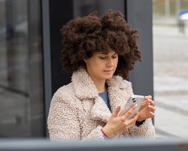 Estilo callejero. Mujer hermosa joven moderna de piel blanca. Con tipo de cabello Afro Hair Style. El peinado exuberante utiliza el teléfono móvil para enviar mensajes de texto. Retrato en la ciudad con el telón de fondo del edificio de hormigón.