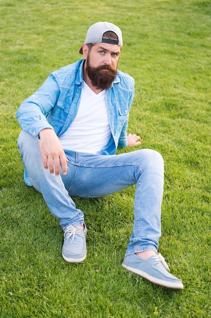 Con estilo y cómodo para vestir. Hipster con estilo en la gorra de béisbol sentado sobre la hierba verde al aire libre. Hombre barbudo con ropa de calle de verano con estilo. Modelo de moda con estilo casual y elegante.